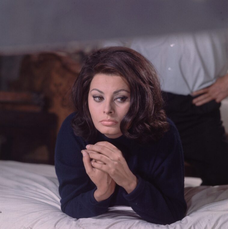 Sophia Loren lays on a bed wearing a blue turtleneck sweater while an unidentified man stands behind her in November 1964, London, England. | Source: Getty Images