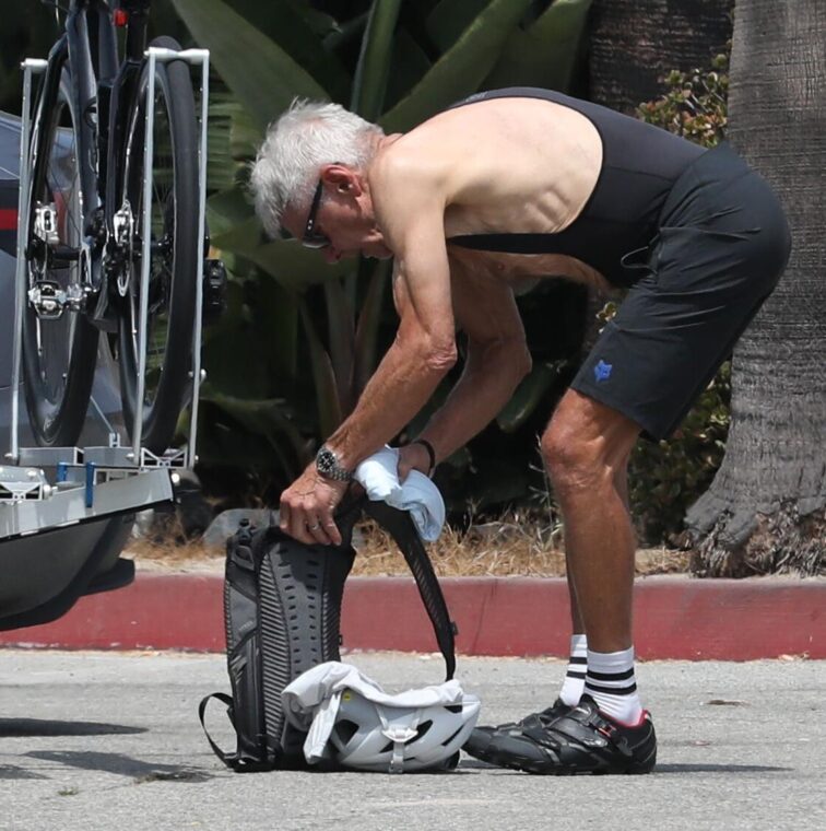 Harrison Ford in cycling attire bending over a backpack and helmet.