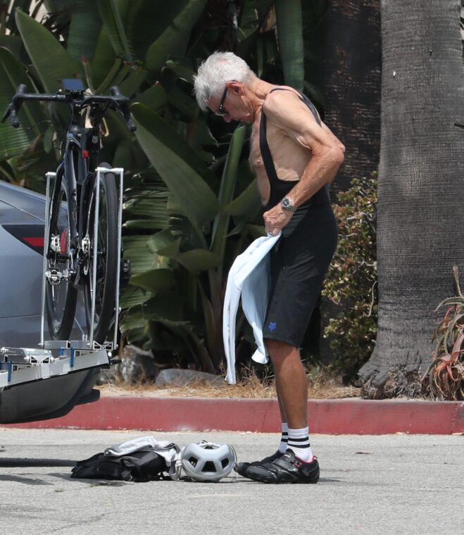Harrison Ford, in cycling gear, stands by his bike, holding a light blue shirt.
