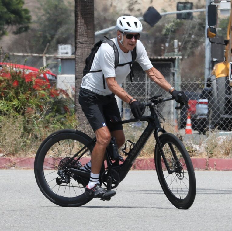 Harrison Ford, in a white long-sleeved shirt and black shorts, walking away from a car with bicycles on a bike rack.