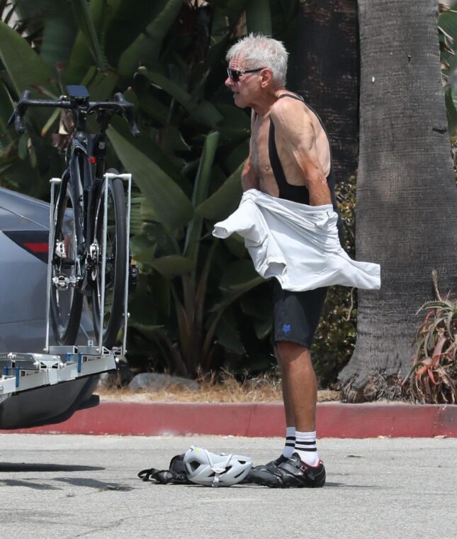 Harrison Ford in cycling gear next to a bike on a rack.